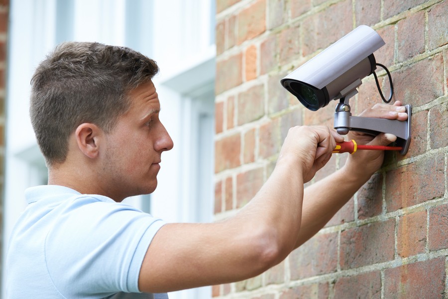 Man installing a security camera on the side of a brick building. 