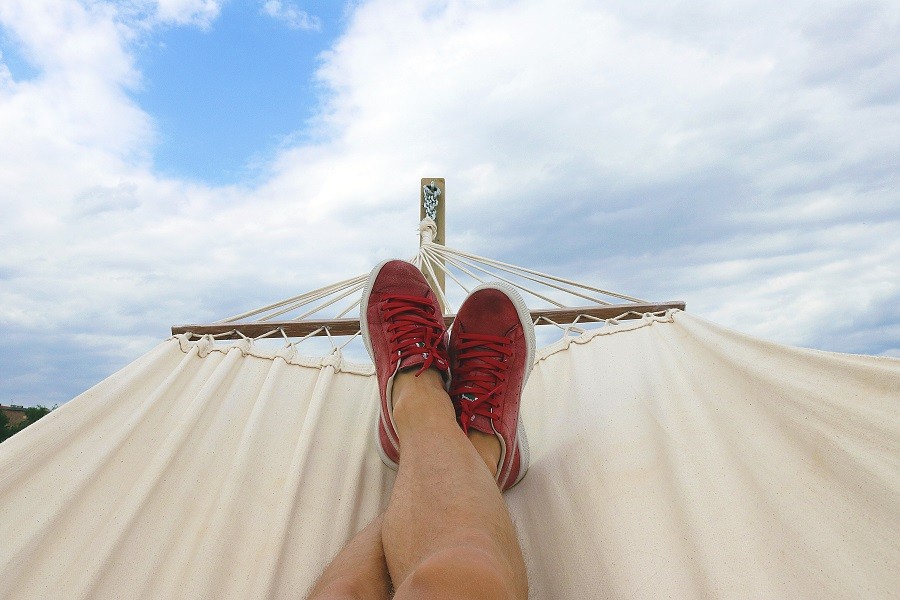 Man's Lower legs crossed on the hammock with sockless red sneakers on his feet and blue sky and fluffy white clouds above him.  