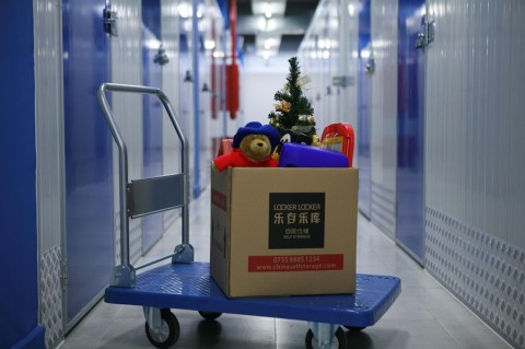 A cardboard box filled with holiday decorations sits on a blue cart in a well-lit self-storage facility with blue and white storage units lining the hallway.